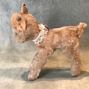 May include: A brown plush toy dog with a white lace collar and a pink flower on the collar. The dog is standing on a grey surface.