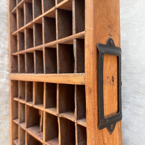 May include: A vintage wooden cubby shelf with numerous small compartments. The wood has a warm, natural tone, and a dark metal label holder is attached to the side. The shelf is designed for organizing small items and has a rustic, aged appearance.