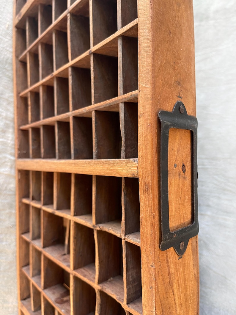 May include: A vintage wooden cubby shelf with numerous small compartments. The wood has a warm, natural tone, and a dark metal label holder is attached to the side. The shelf is designed for organizing small items and has a rustic, aged appearance.