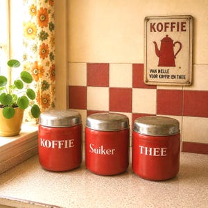 Könnte beinhalten: Drei rote Küchenbehälter mit silbernen Deckeln, beschriftet mit "KOFFIE", "Suiker" und "THEE" in weißer Schrift. Ein Vintage-Schild darüber zeigt "KOFFIE" mit einer Teekannen-Illustration. Der Hintergrund zeigt ein kariertes Fliesenmuster und einen Blumenvorhang.