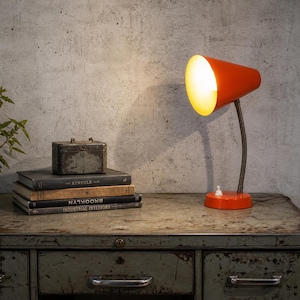 Puede incluir: Una lámpara de escritorio naranja con una pantalla cónica y un cuello flexible de metal oscuro. La lámpara está sobre una base naranja a juego. Junto a la lámpara hay libros apilados y una pequeña caja de metal. Los libros incluyen los títulos "The Kinfolk" e "Industrial Interiors."