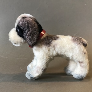 Puede incluir: Un pequeño perro de peluche blanco y negro con un collar rojo. El perro está de pie sobre una superficie gris.