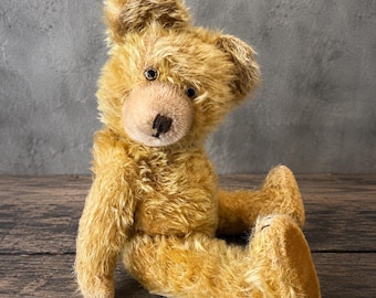 May include: A golden-brown teddy bear with long, shaggy fur, dark round eyes, and a stitched dark brown nose. The bear is in a seated position with its head tilted slightly to the side, featuring a lighter-colored muzzle and textured paw pads.