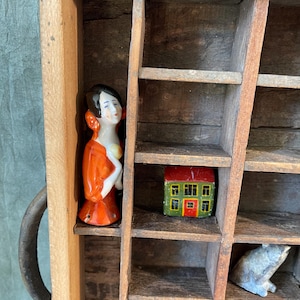 May include: A vintage wooden printer's tray with multiple compartments, containing small ceramic figurines. The figurines include a woman in an orange dress, a miniature house with a red roof, and a small animal. The tray has a metal handle.