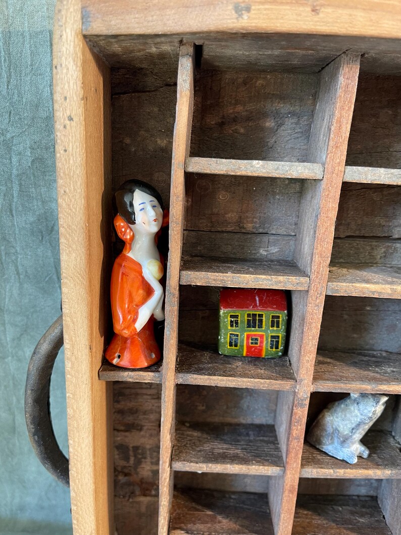 May include: A vintage wooden printer's tray with multiple compartments, containing small ceramic figurines. The figurines include a woman in an orange dress, a miniature house with a red roof, and a small animal. The tray has a metal handle.