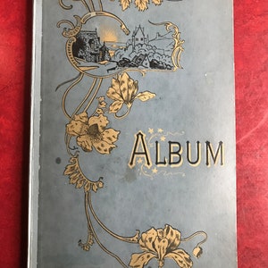 May include: A vintage blue and gold photo album with an ornate floral design and the word "ALBUM" printed in gold letters. The cover features a small illustration of a seaside village.