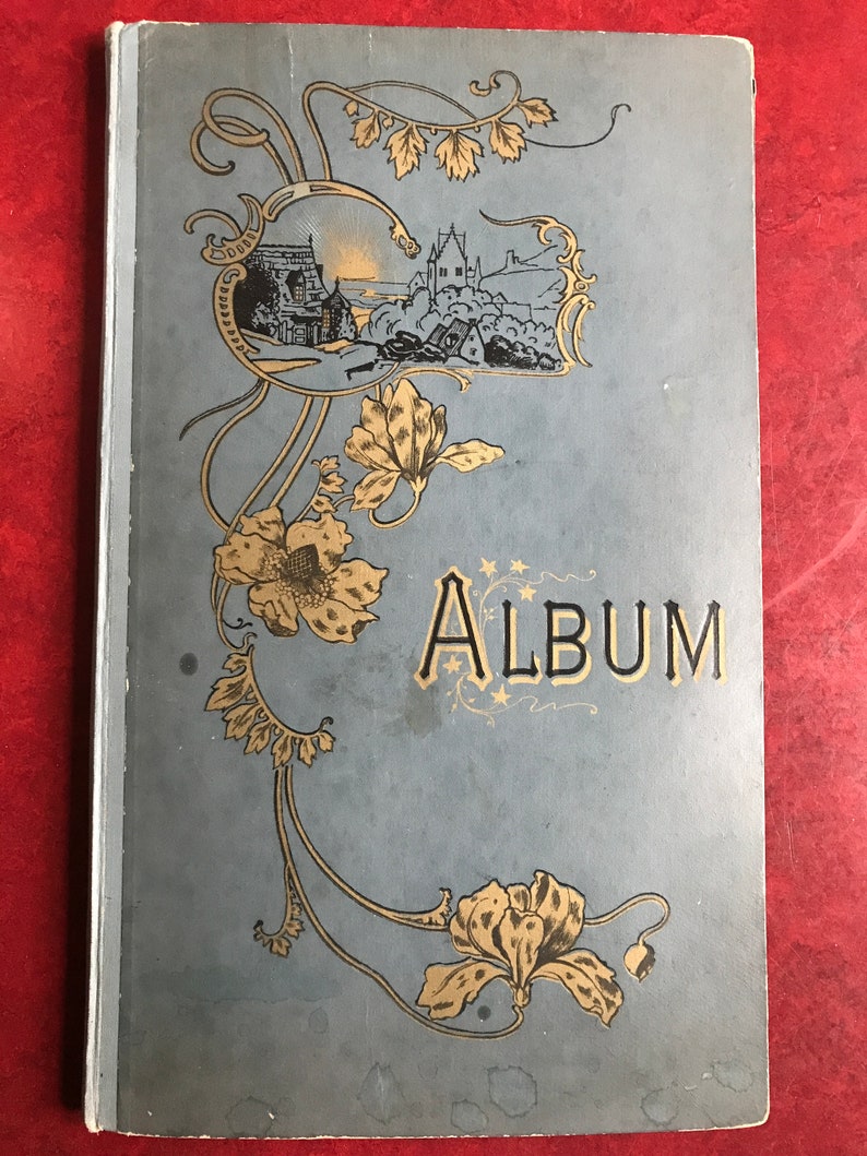 May include: A vintage blue and gold photo album with an ornate floral design and the word "ALBUM" printed in gold letters. The cover features a small illustration of a seaside village.