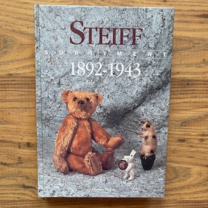 May include: A hardcover book titled Steiff Sortiment 1892-1943 with a gray textured cover. The front features a large brown teddy bear, a small white bear holding a circular object, and a small pig figurine standing on a black base.