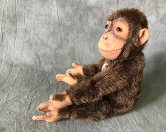 Puede incluir: Un juguete de mono de peluche marrón con grandes ojos marrones y una sonrisa cosida. El mono está sentado con las piernas cruzadas y los brazos extendidos.