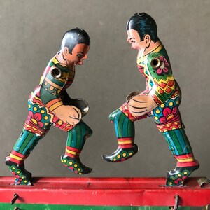 May include: Two colorful tin toy soccer players in mid-kick, with a red and green base. The players are wearing green, blue, yellow, and red uniforms with floral patterns.