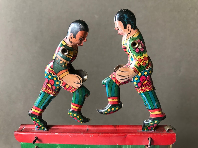 May include: Two colorful tin toy soccer players in mid-kick, with a red and green base. The players are wearing green, blue, yellow, and red uniforms with floral patterns.