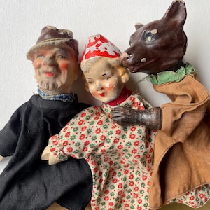 May include: Three vintage hand puppets featuring a male figure in a black outfit, a child figure in a red and white floral dress with a matching hat, and a brown wolf figure wearing a tan tunic with a green collar, all with painted faces.