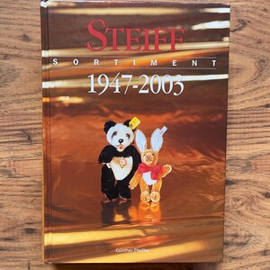 May include: A hardcover reference book titled Steiff Sortiment 1947-2003 by Günther Pfeiffer. The cover features a brown background with a black and white panda bear plush toy and a light brown rabbit plush toy wearing a red bow tie.