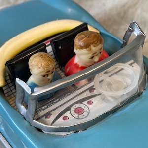 Könnte beinhalten: Ein blaues Spielzeugauto mit einer gelben Banane auf dem Dach. Zwei Figuren mit bemalten Gesichtern sitzen im Auto. Das Auto hat eine silberne Windschutzscheibe und ein weißes Lenkrad.