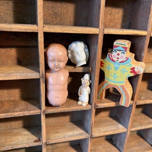 May include: A wooden display case with multiple compartments, each holding a different antique figurine. The figurines include a baby doll, a white bust, a small bear figure, and a colorful wooden clown. The case is made of aged wood.