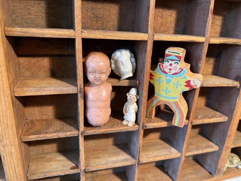 May include: A wooden display case with multiple compartments, each holding a different antique figurine. The figurines include a baby doll, a white bust, a small bear figure, and a colorful wooden clown. The case is made of aged wood.