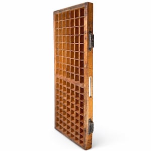 May include: A rectangular wooden letterpress type case featuring a grid of numerous small square compartments. The frame is constructed from light brown wood and includes two dark metal side handles and a vertical slot on the side edge.