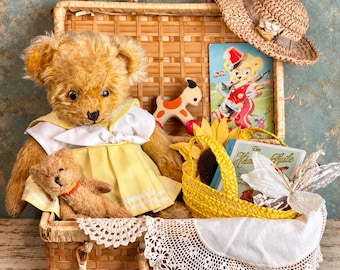 Puede incluir: Una maleta de mimbre tejida que contiene un oso de peluche grande de color marrón dorado con un vestido amarillo, un oso de peluche pequeño, un sombrero de paja, un juguete de madera, una cesta amarilla, un libro titulado Die Hansi-Fute y un tapete de encaje.
