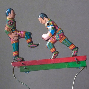 May include: Two colorful tin toy figures of soccer players on a red and green platform. The figures are painted with bright colors and intricate details. The players are in mid-kick and appear to be playing a game of soccer.