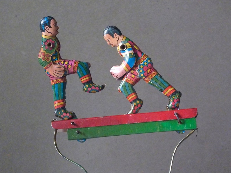 May include: Two colorful tin toy figures of soccer players on a red and green platform. The figures are painted with bright colors and intricate details. The players are in mid-kick and appear to be playing a game of soccer.