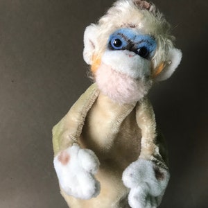 Puede incluir: Un títere de mono de peluche con pelaje blanco, ojos azules y una cara marrón claro. El mono lleva una túnica de tela verde claro y marrón.