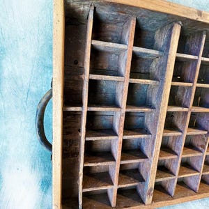 May include: A vintage wooden printer's tray with multiple small compartments. The tray is made of aged wood with a natural finish and a metal handle. The compartments are arranged in a grid pattern, ideal for organizing small items.