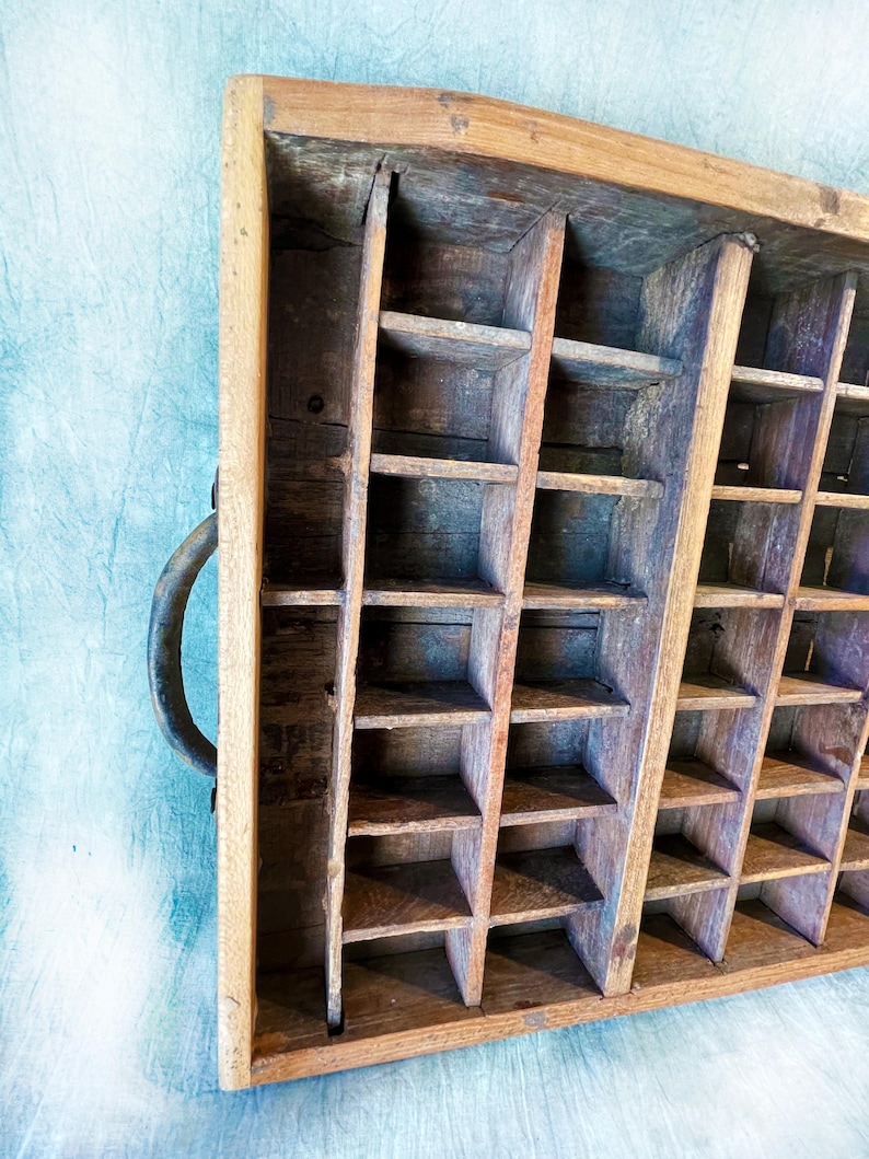 May include: A vintage wooden printer's tray with multiple small compartments. The tray is made of aged wood with a natural finish and a metal handle. The compartments are arranged in a grid pattern, ideal for organizing small items.