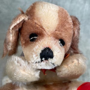 Puede incluir: Un juguete de peluche vintage con forma de perro con pelaje marrón y crema. El perro tiene ojos negros, una nariz negra y un lazo rojo. El juguete es una representación clásica de un cachorro.