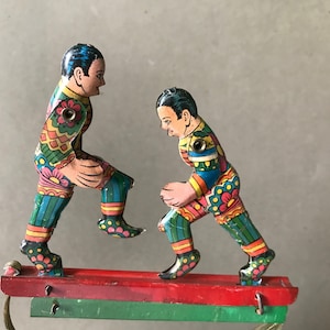 May include: Two colorful tin toy figures of people playing a game. The figures are painted in bright colors with floral patterns and are standing on a wooden platform.