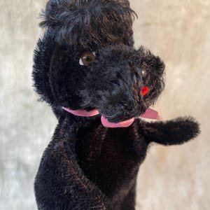 May include: A vintage black poodle plush toy with a pink ribbon around its neck. The plush has brown eyes and a red nose. The poodle is made of a soft, fuzzy material.