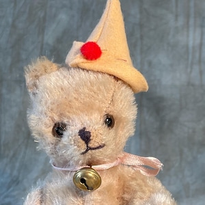 May include: A vintage teddy bear with light brown fur, wearing a cream-coloured hat with a red pom-pom and a pink ribbon around its neck. A small gold bell hangs from the ribbon. The bear has dark eyes and a stitched nose.