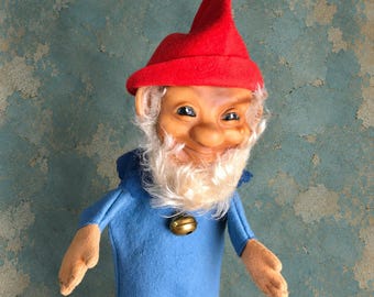 May include: A hand puppet featuring a gnome with a bright red felt hat, a tan face, and a white synthetic beard. The gnome wears a blue felt tunic with a small, round, metallic gold-colored bell attached at the neckline, centered on the chest.