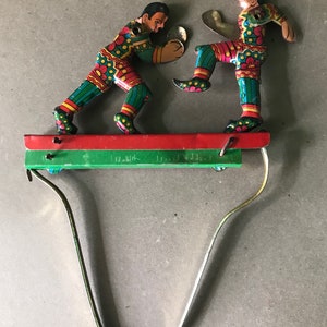 May include: A vintage tin toy of two figures boxing on a platform with a spring-loaded mechanism. The figures are painted in bright colors with green, red, and yellow accents. The figures are wearing colorful clothing and have exaggerated features.