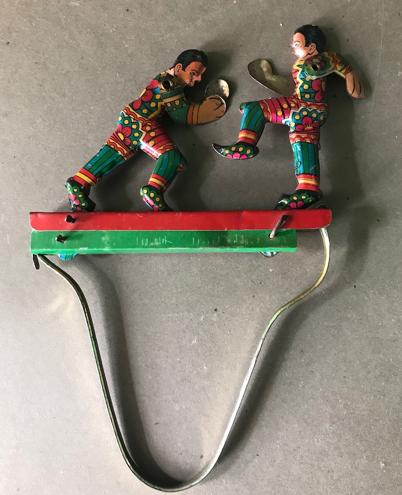 May include: A vintage tin toy of two figures boxing on a platform with a spring-loaded mechanism. The figures are painted in bright colors with green, red, and yellow accents. The figures are wearing colorful clothing and have exaggerated features.