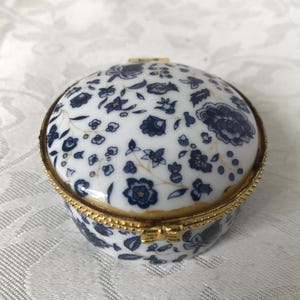 Puede incluir: Un joyero redondo de porcelana con base blanca, estampado floral azul oscuro y borde de metal dorado. La tapa abovedada está decorada con flores azules detalladas y finas líneas doradas, asegurada por un pequeño cierre de metal dorado.