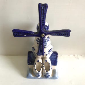 May include: A ceramic windmill figurine in blue and white, featuring a traditional Dutch design. The windmill has a blue roof, blades, and decorative elements. Two white clogs with blue floral patterns are at the base. The figurine is a decorative item.