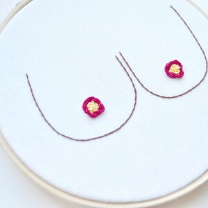 May include: Embroidered hoop art featuring two stylized breasts with pink and yellow flowers at the center.