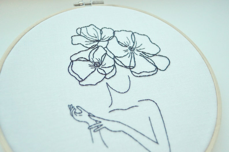 K&ouml;nnte beinhalten: Schwarz-wei&szlig;-Stickerei einer Person mit Blumen auf dem Kopf. Die Person ist mit einer einfachen Umrandung ihres K&ouml;rpers und ihrer H&auml;nde dargestellt. Die Blumen sind stilisiert und haben ein einfaches Design.