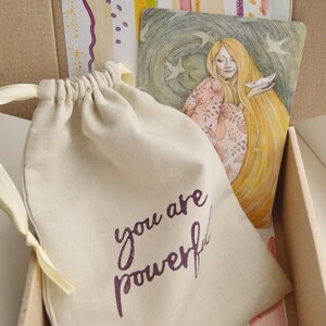 May include: A small, beige drawstring bag with the words "you are powerful" printed in purple. The bag is sitting in a cardboard box with a colorful patterned paper lining.