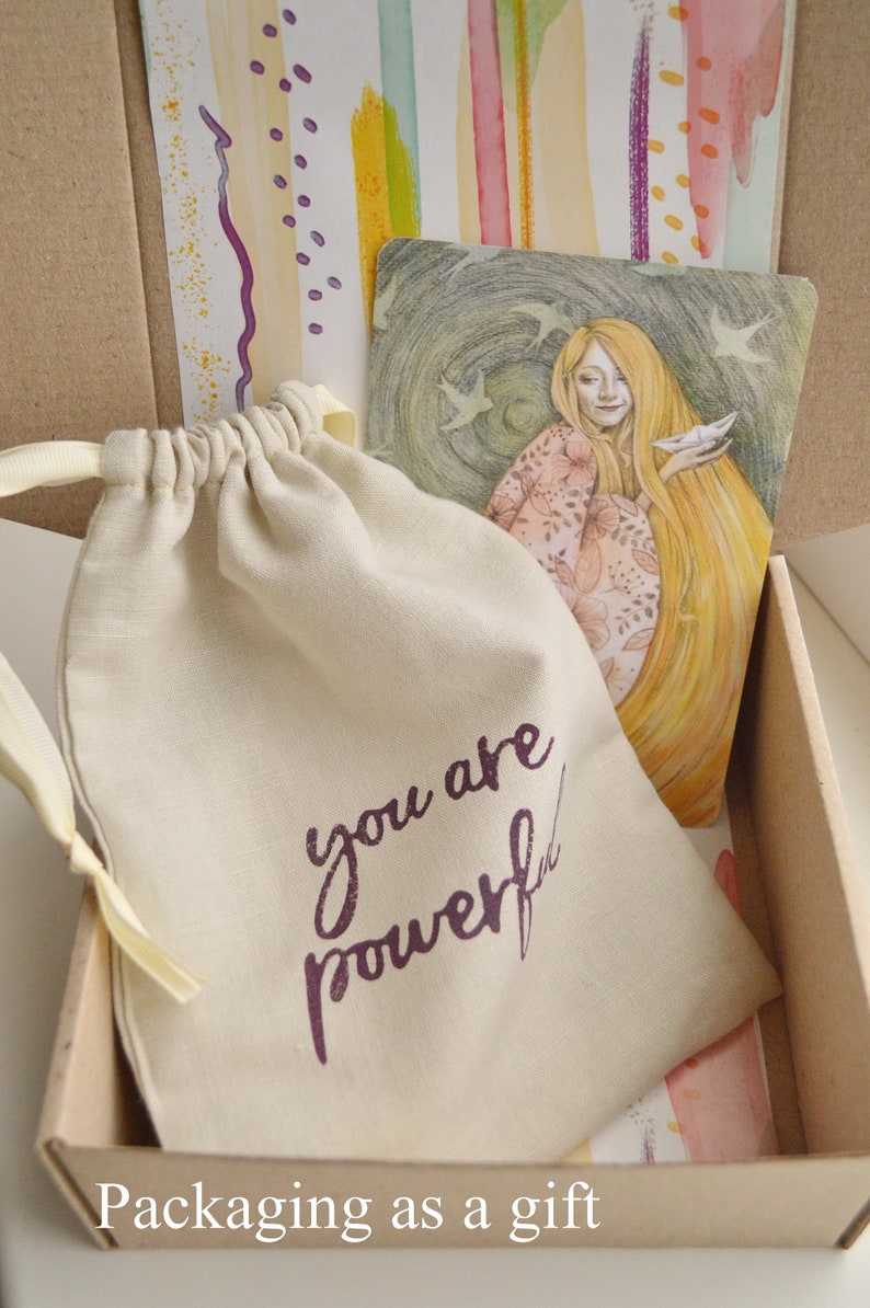 May include: A small, beige drawstring bag with the words "you are powerful" printed in purple. The bag is sitting in a cardboard box with a colorful patterned paper lining.
