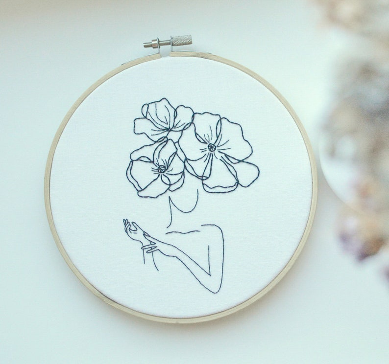 K&ouml;nnte beinhalten: Stickerei auf einem Stickrahmen mit einer Linienzeichnung einer Person mit Blumen als Kopf. Die Blumen sind stilisiert und haben jeweils f&uuml;nf Bl&uuml;tenbl&auml;tter.