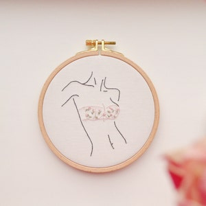 May include: A white embroidery hoop with a black line drawing of a person wearing a floral patterned bra. The hoop is made of wood and has a gold metal clasp.