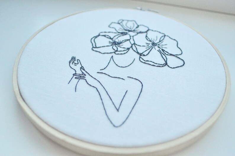 K&ouml;nnte beinhalten: Schwarz-wei&szlig; gestickte Bilderrahmenkunst mit einer Linienzeichnung einer Person mit Blumen als Kopf.