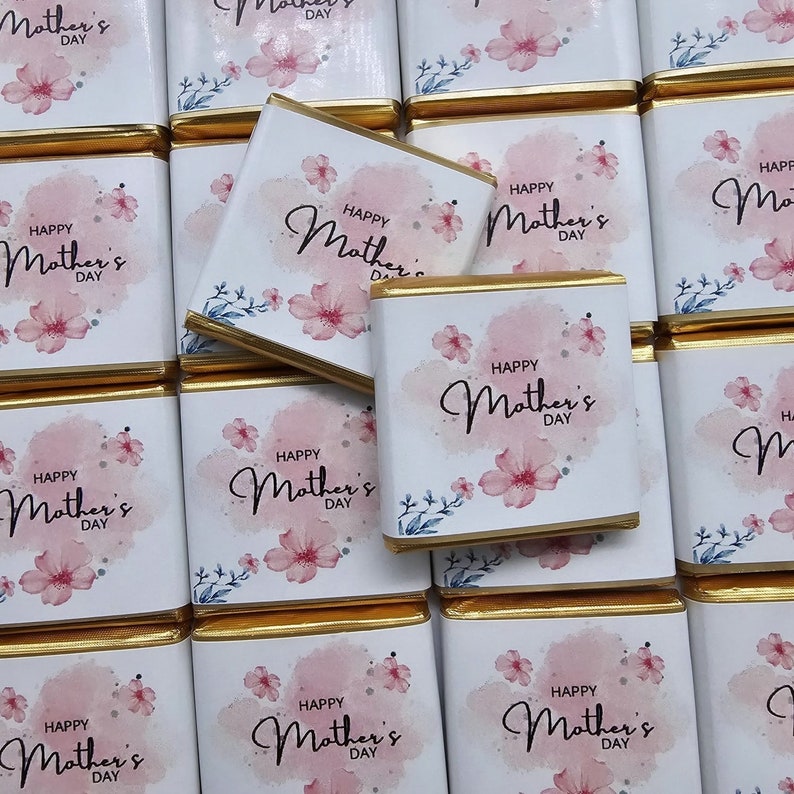 Pu&ograve; includere: Primo piano di diverse piccole barrette di cioccolato avvolte in carta bianca con un bordo dorato. Ogni barretta presenta un disegno ad acquerello di fiori rosa e il testo "Happy Mother's Day".