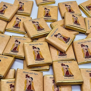 May include: A stack of small, square, brown chocolate bars with gold foil wrappers. Each bar has a design of a couple in traditional clothing and the text "Ahsan and Aqsa" with the date "22nd June 2024".