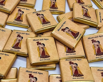 May include: A stack of small, square, brown chocolate bars with gold foil wrappers. Each bar has a design of a couple in traditional clothing and the text "Ahsan and Aqsa" with the date "22nd June 2024".