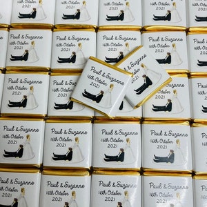 May include: A collection of small, gold-wrapped chocolate bars with white labels. Each label features a cartoon illustration of a bride and groom, along with the text "Paul & Suzanne" and "16th October 2021".