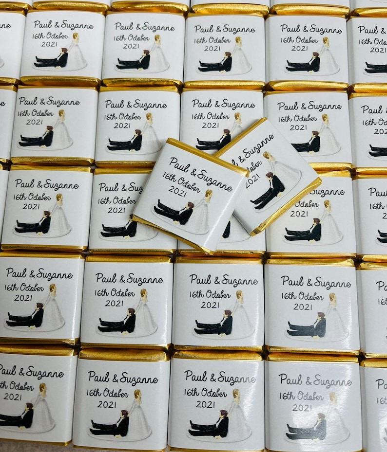 May include: A collection of small, gold-wrapped chocolate bars with white labels. Each label features a cartoon illustration of a bride and groom, along with the text "Paul & Suzanne" and "16th October 2021".