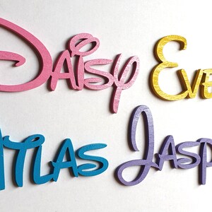 May include: Four wooden names in a Disney font style. The names are Daisy in pink, Evelyn in gold, Atlas in blue, and Jasper in purple.