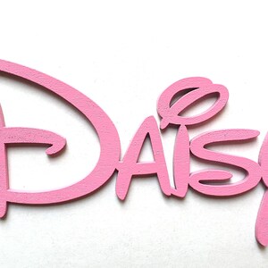May include: Pink wooden cutout of the name "Daisy" in a stylised font reminiscent of the Disney logo.